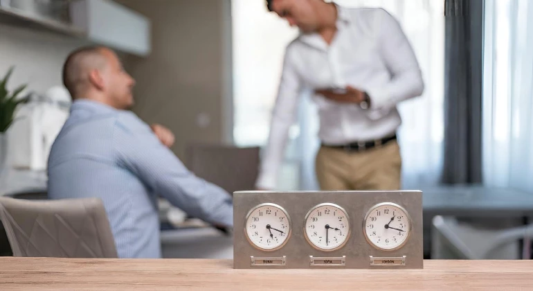 Tres relojes colocados sobre una mesa de oficina. Al fondo, dos personas conversan en un entorno corporativo. La imagen transmite la importancia de la gestión del tiempo en contextos internacionales, en sintonía con los plazos legales para reclamar vicios ocultos